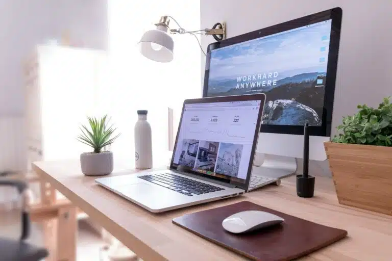 Portátil y monitor sobre una mesa de madera con una planta, destacando la ergonomía sostenible.